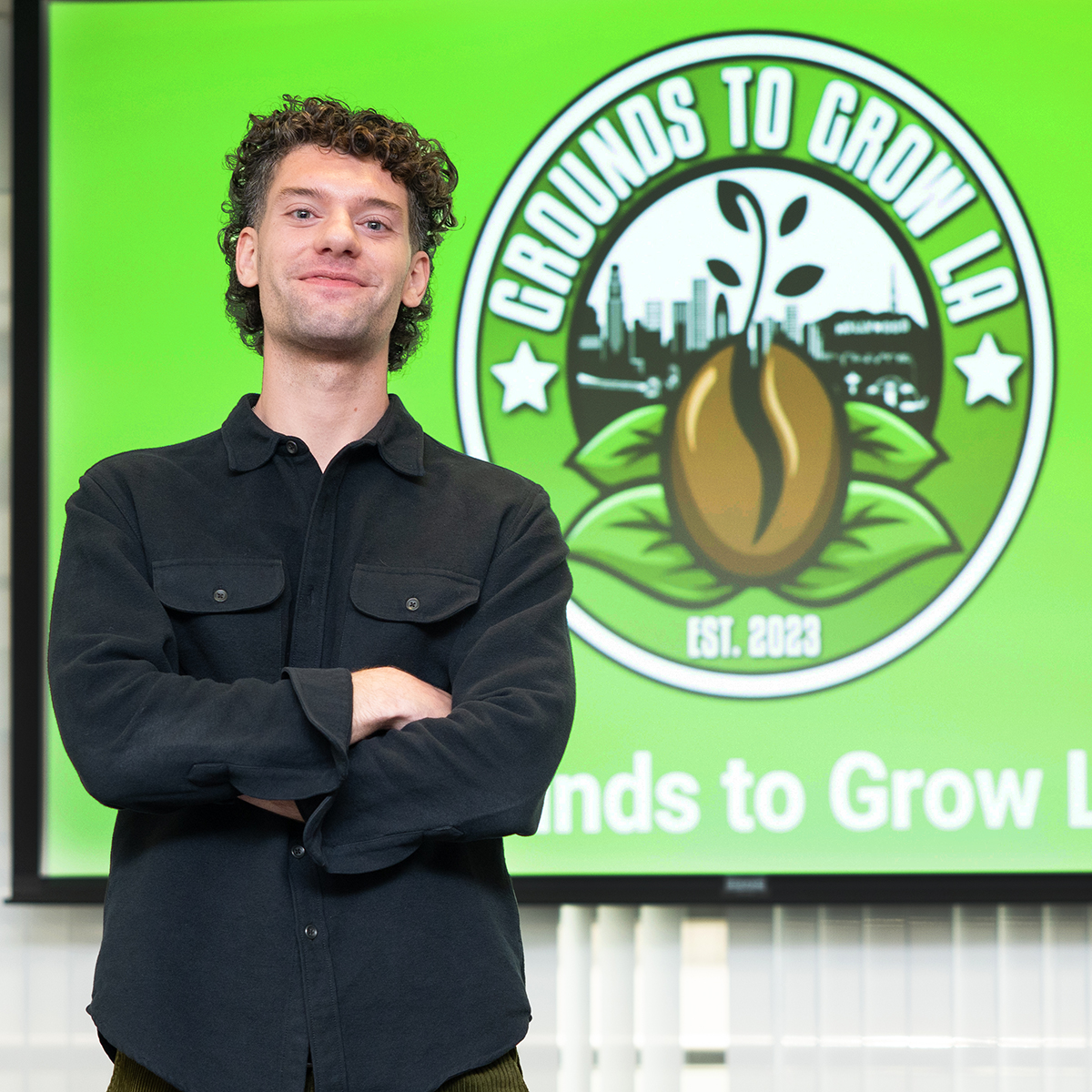 A student standing with arms crossed in front of a projection screen displaying the logo for 